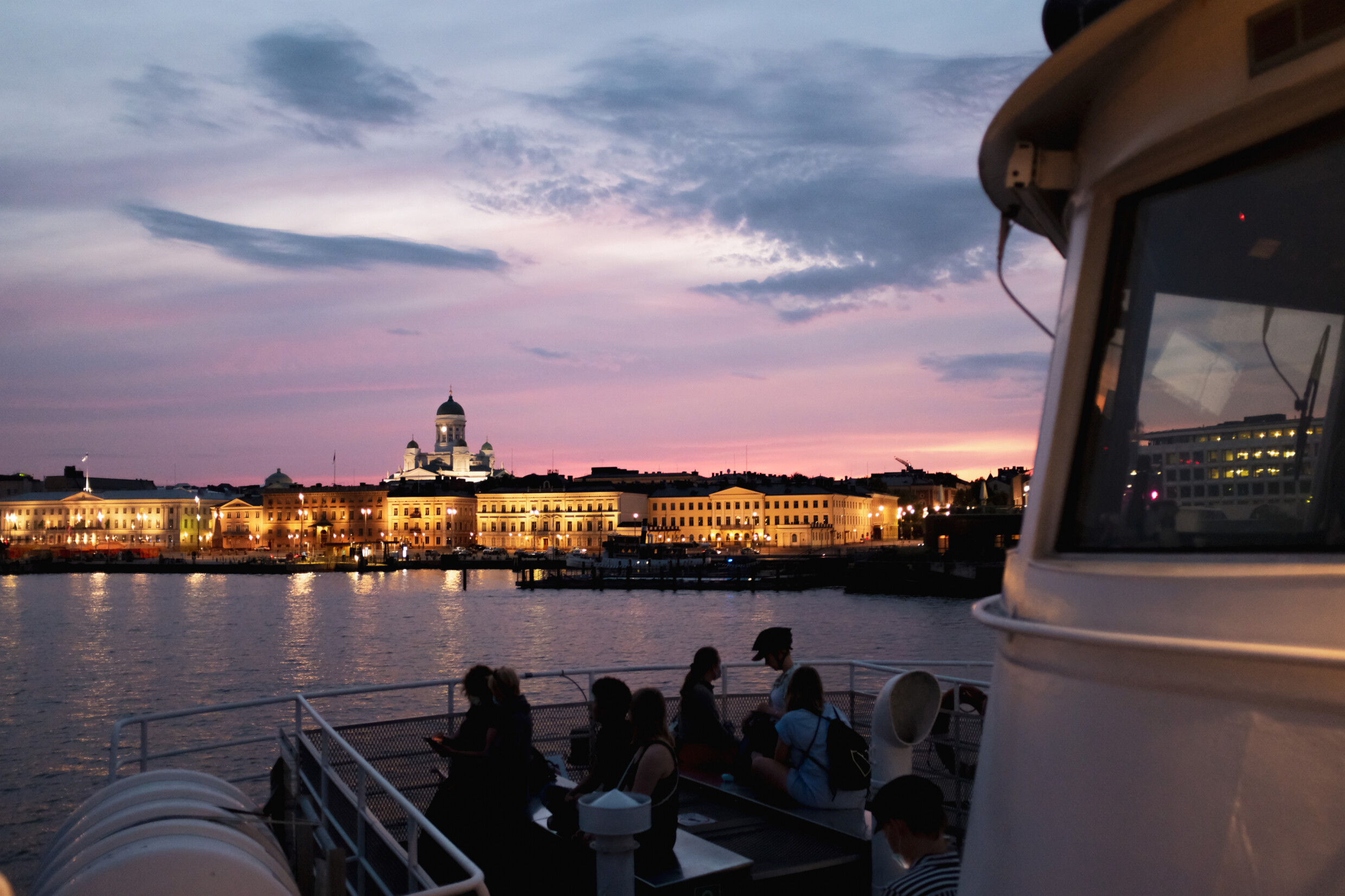 Suomenlinnan lautta jonka kannella ihmisiä, tustalla tuomiokirkko auringonlaskussa.